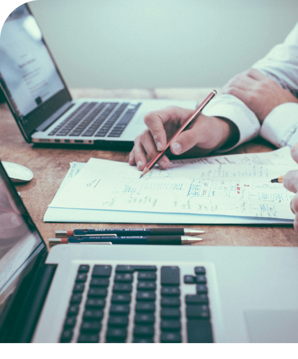 Someone sits at a table with 2 laptops open on it, the person is holding a pen