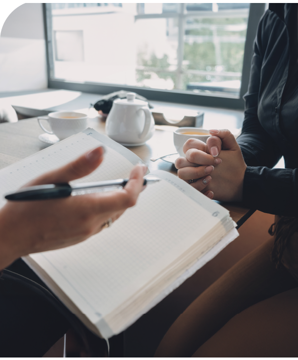 A woman sits at table with her hands folded in front of someone else holding a pen and paper.