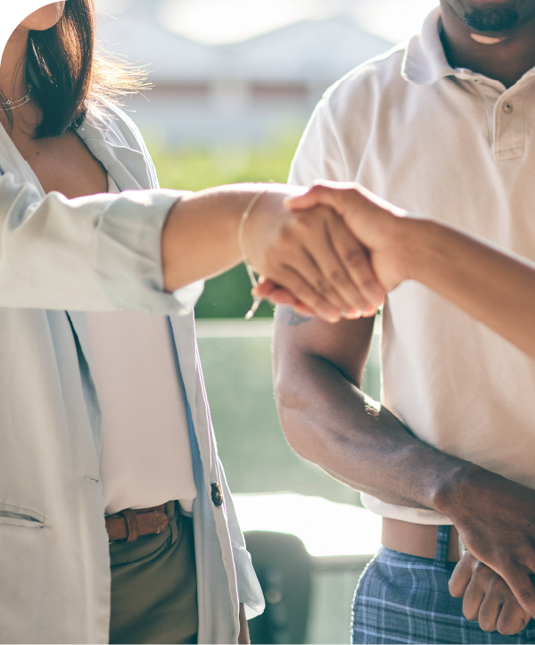 A man and woman stand in front of someone out of frame. The woman shakes hands with the third person.
